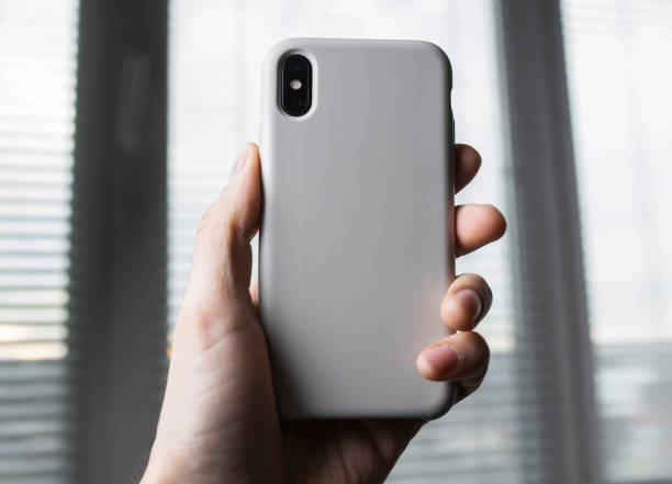 istock Smart phone on the blurred background of the winwow in a white plastic case back view. Smart phone in man's hand. Template of phone case 1293876411