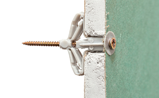 Grey plastic butterfly-type dowel fixed in pre-drilled hole in sheet of moisture-resistant drywall isolated on white background