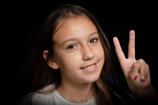 Close up portrait of a cheerful little girl making the victory gesture with her hand stained in red paint on black studio background. Joy concept
