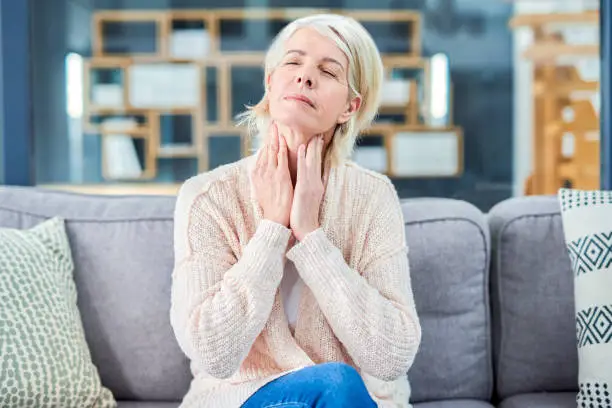 Shot of a senior woman suffering from throat pain while sitting on the sofa at home