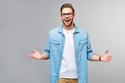 young man wearing jeans shirt welcoming you with a smile on his face and his arms wide open standing over grey background.