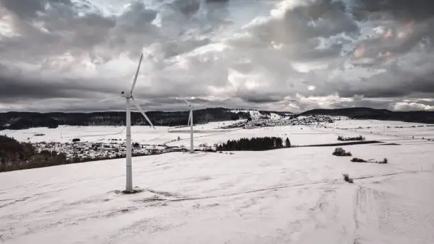 Wind Turbines in white snow covered rural Winter Landscape under partly sunny dramatic winter skyscape. Green Renewable Energy, Alternative Energy Environment Concept Shot. Drone Point of View. Baden Württemberg, Germany, Europe