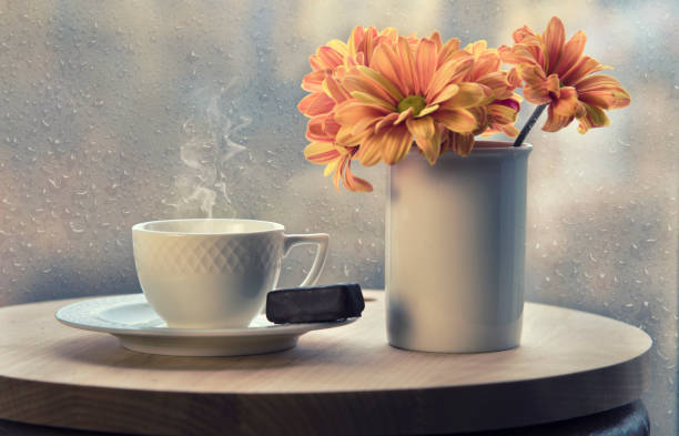 White coffee cup with smoke, vase with orange flowers on table in cafe White coffee cup with smoke, vase with orange flowers on table in cafe. Raining outside. Late autumn and coziness concept. Rain drops on window glass. Selective focus. rainy cafe stock pictures, royalty-free photos & images