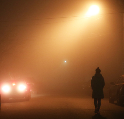 The silhouette of a girl on a dark night and foggy street is illuminated by a yellow street lamp. The fog is very dense, everything is blurry