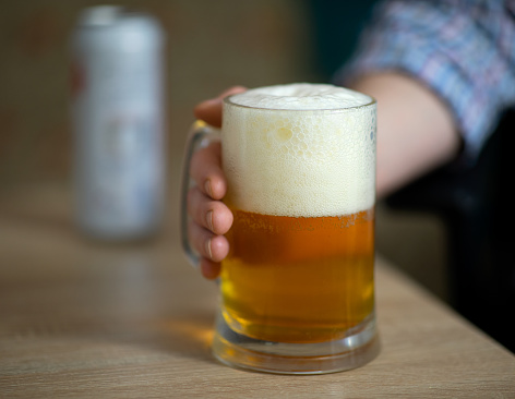Male hand holding large mug full of cold fresh alcoholic beer on wooden background, man at home, selective focus.