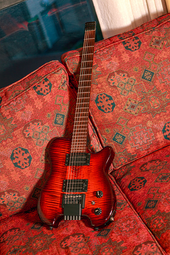 Bogota, Colombia - A headless electric guitar displayed in a home interior, on a sofa that is furbished with a rich, design woven, red fabric. The tuning pegs can be seen at the bottom of the guitar, at the exact opposite end of where one would usually find them. Horizontal format; no people. Copy Space. Note to Inspector: Logos and other brand markings have been removed.