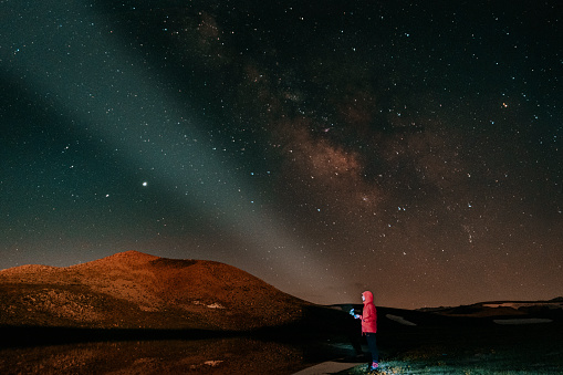 Male tourist have a rest in his camp near the at night. Man standing near campfire and tent under beautiful night sky full of stars and milky way, and enjoying night scene. Panoramic landscape