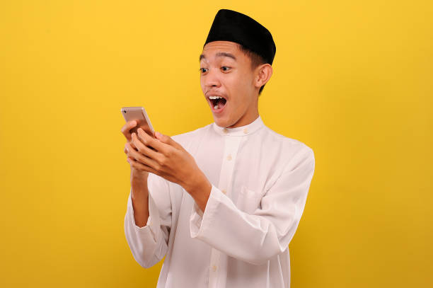 ハッピーショック若いアジアのイスラム教徒の男は携帯電話の画面を見て携帯電話を保持しているイスラム教徒の服を着て - 3656 ストックフォトと画像