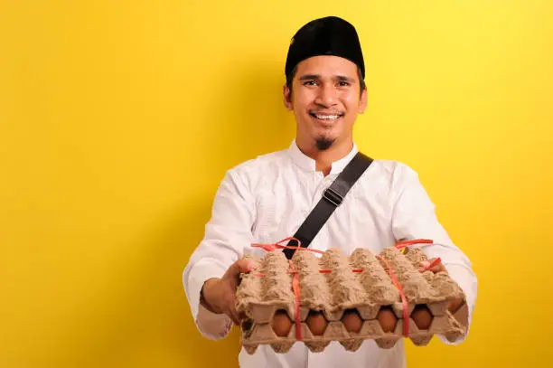 In Ramadan Kareem, Smiling Happy Young Asian Muslim man bring  a plate of eggs to give for everyone need, isolated on yellow background