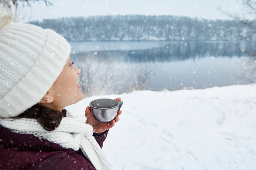 A woman in white knitted hat and scarf looking away and enjoying beautiful snowy day while drinking hot tea on the background of a snow-covered lake