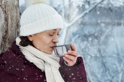 Woman in white knitted woolen hat enjoying beautiful snowy day while drinking hot tea. Falling snow