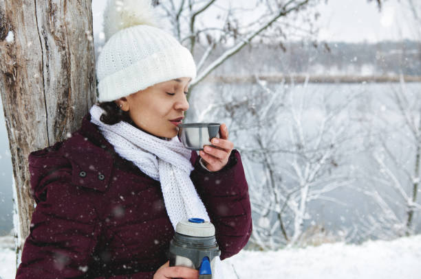 istock Woman in white knitted woolen hat enjoying beautiful snowy day while drinking hot tea. Falling snow 1293995961