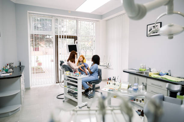istock Modern dentist's office. Mother and little daughter having a regular check up and talking with their dentist, young African woman. Oral care, caries prevention 1294004872