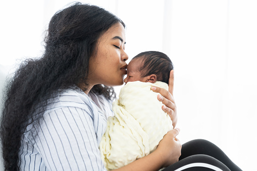 Happy African American mother kissing her newborn baby while sitting on bed. Mom carrying her afro infant child on hands with kindly. family, love, happy and new life concept