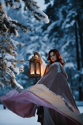 Epic fantasy shot: Young woman spinning her flying cloak holding vintage lantern in snowy fir and pine forest at night. Medieval fairytale cosplay concept