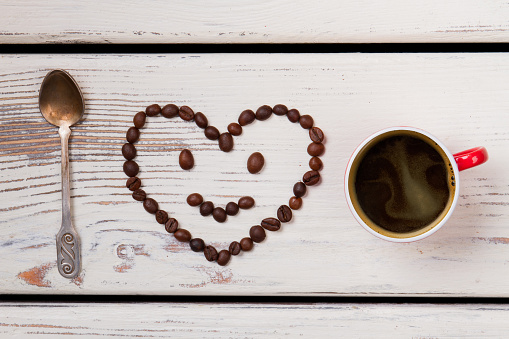 Cup of hot coffee and heart-shaped smiley and teaspoon. White wood background. Top view flat lay.