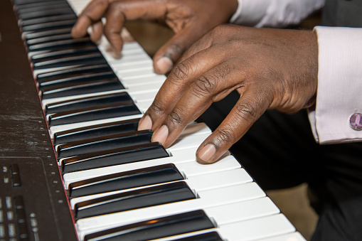 Fingers of an old African American man playing a keyboard piano wearing a black suit and pink shirt
