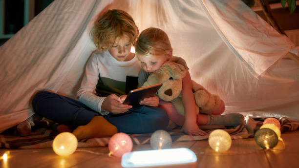 istock Adorable little children, boy and girl using digital tablet while spending time together, sitting in a hut made with bedsheets at home 1294041251