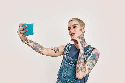 A white tattooed pierced woman wearing denim overall making a selfie on her smartphone smiling with her hand near her face on a white background