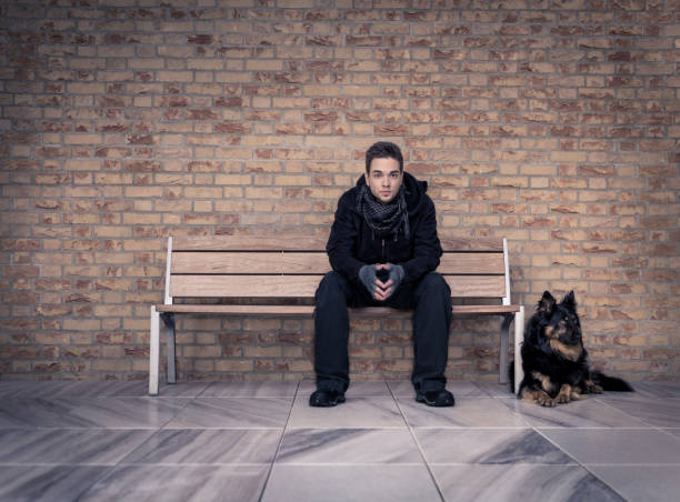 A young man, with his black dog next to him, sitting on a modern bench in a room with a marble floor and brick walls. The man is wearing black clothes, knitted fingerless gloves and a gray shawl.