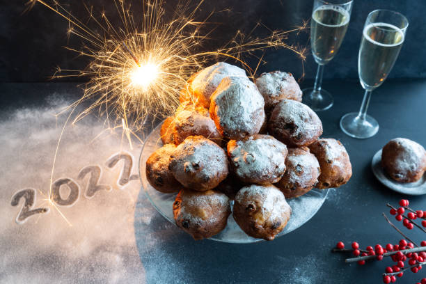 gelukkig nieuwjaar met het jaar 2022 geschreven in poedersuiker naast een kom oliebollen met glitters en champagne - oliebollen stockfoto's en -beelden
