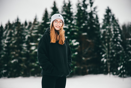 Happy smiling teenage girl standing in front of snow covered winter landscape of Black Forest in Germany, looking over to the camera with a bright smile. Millennial Generation Real People Winter Outdoor Portrait.