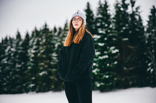 Teenage Woman standing in front of snow covered winter landscape of Black Forest in Germany, looking over to the camera with a straight cool attitude. Millennial Generation Real People Winter Outdoor Portrait.