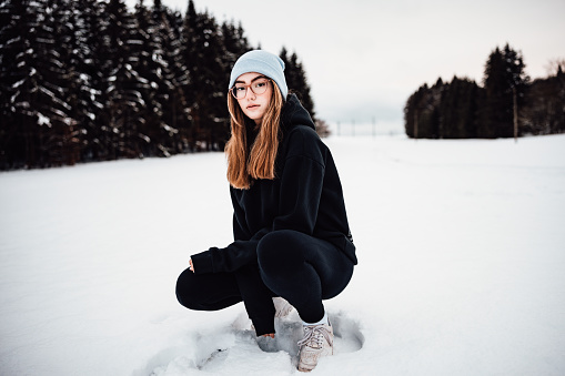 Fashionable Teenage Girl kneeling in the snow in cold german Black Forest Winter Landscape. Looking over towards the camera with a cool attitude and a slight smile. Millennial Generation Real People Winter Outdoor Portrait.