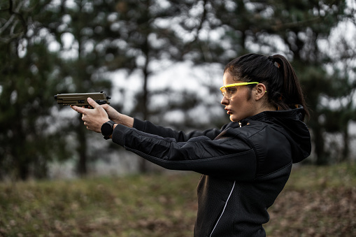 Beautiful young woman wearing hooded shirt and protective eyeglasses while aiming and practising shooting from pistol at shooting range
