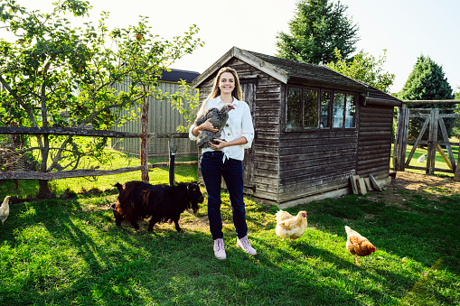 Caucasian woman in early 20s smiling at camera while standing in enclosed grass area with grazing free range chickens and goat.