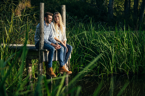 Caucasian couple in early 20s wearing casual clothing and talking as they sit at end of small dock overlooking pond surrounded by tall grasses.