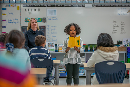 An adorable African American elementary student talks about her artwork to her classmates. She stands at the front of the class.