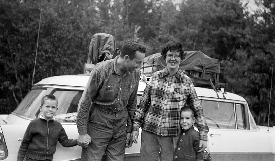 Car packed and ready to go in background for family vacation in 1957, Family of parents and two boys in front.