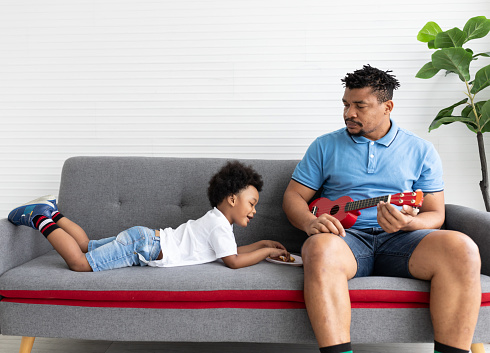Adult African American father sitting on sofa and looking to his little son while lying on sofa and eating cookie. African American father playing with his little son. Happy family and people concept