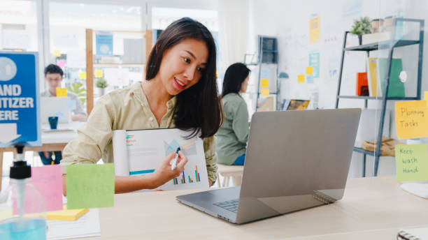 Asia businesswoman social distancing in new normal situation for virus prevention while using laptop presentation to colleagues about plan in video call while work in office.