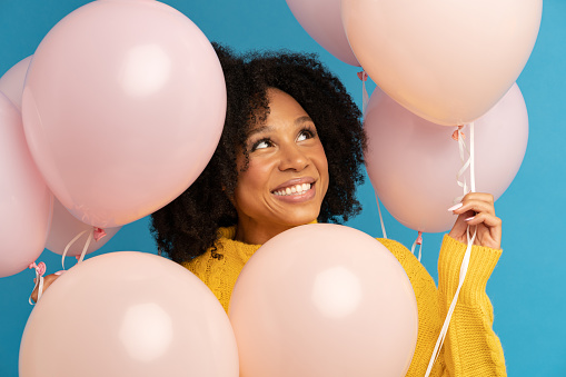 Close up of happy dark skinned woman among pastel pink air balloons with joy and happiness, celebrates birthday, looking up, wears yellow jumper, standing over studio blue background. Festive event.