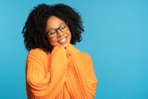 Stylish happy dark skinned millennial woman with curly hair wear orange knitted jumper, holds hands under chin, copy space for advertising, isolated on studio blue background. Winter warm clothes.