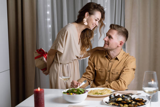 jovem casal na mesa, uma mulher curvada sobre um homem segurando uma caixa com um presente em suas mãos. conceito de celebração do dia dos namorados, aniversário, - men giving balloon women - fotografias e filmes do acervo