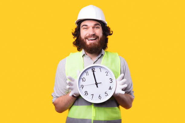 istock Happy young bearded engineer is holding a big rounded clock and smiling is looking at the camera near a yellow wall 1294125711