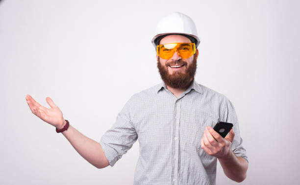 istock A bearded young engineer is holding a smartphone and smiling at the camera is wearing a helmet and protective glasses near a white wall 1294125879