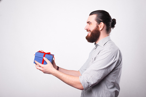 Bearded happy man is holding a gift trying to give it to someone is smiling near a white wall .