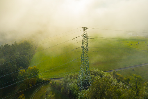 Aerial view of the high voltage power lines and high voltage electric transmission on the terrain in clouds surrounded by trees at sunlight.