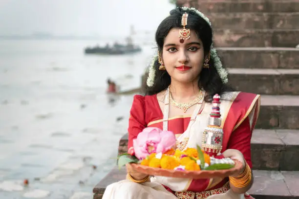 Portrait of beautiful Indian girl in front of ganga river wearing traditional Indian saree, gold jewellery and bangles holding plate of religious offering.