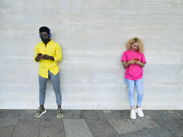 スマートフォンで音楽を聴きながら社会的距離を持つ街の若い多民族の友人 - 保護フェイスマスクを着用した若者 - couple blond hair social gathering women ストックフォトと画像
