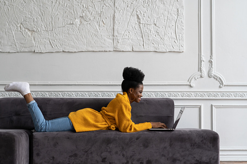 Smiling Afro-American biracial woman with afro hairstyle in yellow cardigan lying on couch, resting, looking at camera webcam and talking on video chat with her friends, watching webinar.