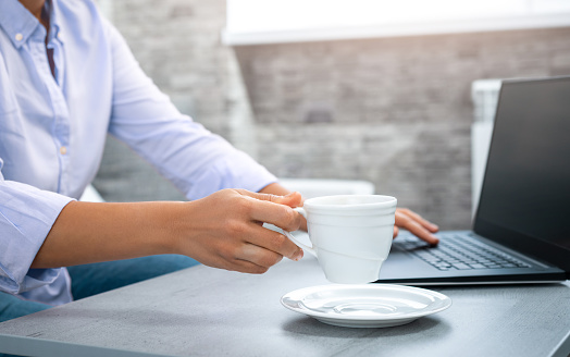 The view of a woman's hand raising a cup with a laptop in the background. laptop. Remote work from home.