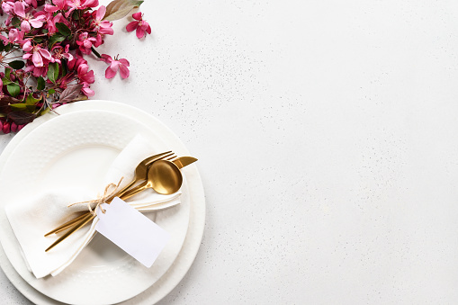 Spring elegance table setting with apple tree flowers, golden cutlery and tag on white table. View from above. Space for text.
