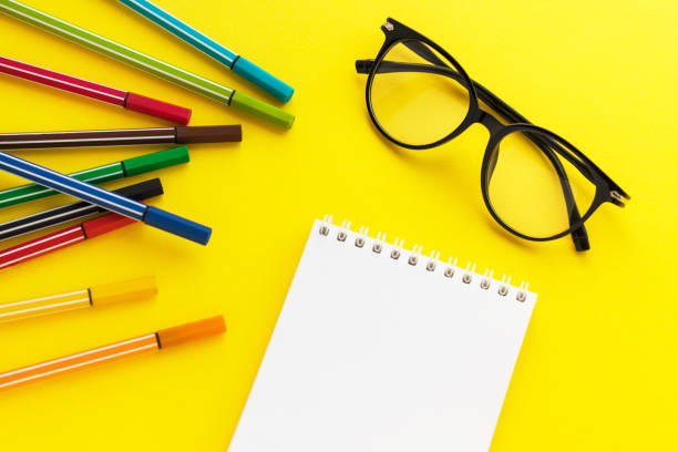 istock Eyeglasses, Blank notepad and colored felt-tip pens on yellow background. Multicolored markers for kid drawing. Flat lay, top view, copy space. 1294189323