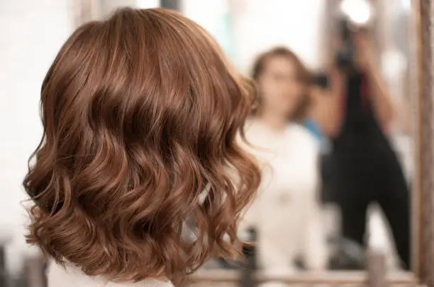 Beautiful wavy hair styling on a young woman with medium brown hair indoors, view from the back with reflection in the mirror, close-up