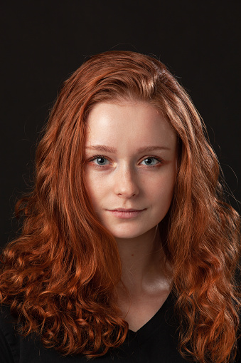 Close up studio portrait of 18 year old woman with long curly red hair on black background